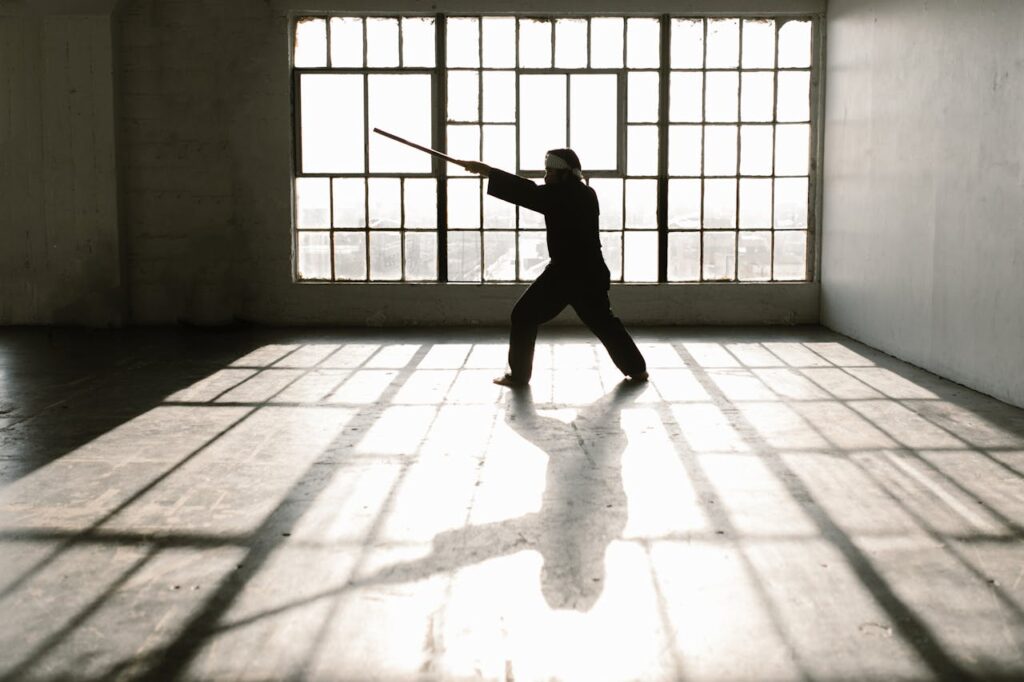 pexels-photo-7045444 Silhouette of martial artist practicing with a staff in a sunlit studio.
